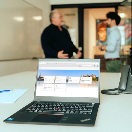 Ein Laptop, auf dem die neue E-Akte geöffnet ist. Im Hintergrund unterhalten sich zwei Männer.